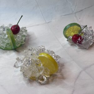 Three faux drink garnishes featuring cherries, lemon or lime slices, and sparkling ice pieces on marble.