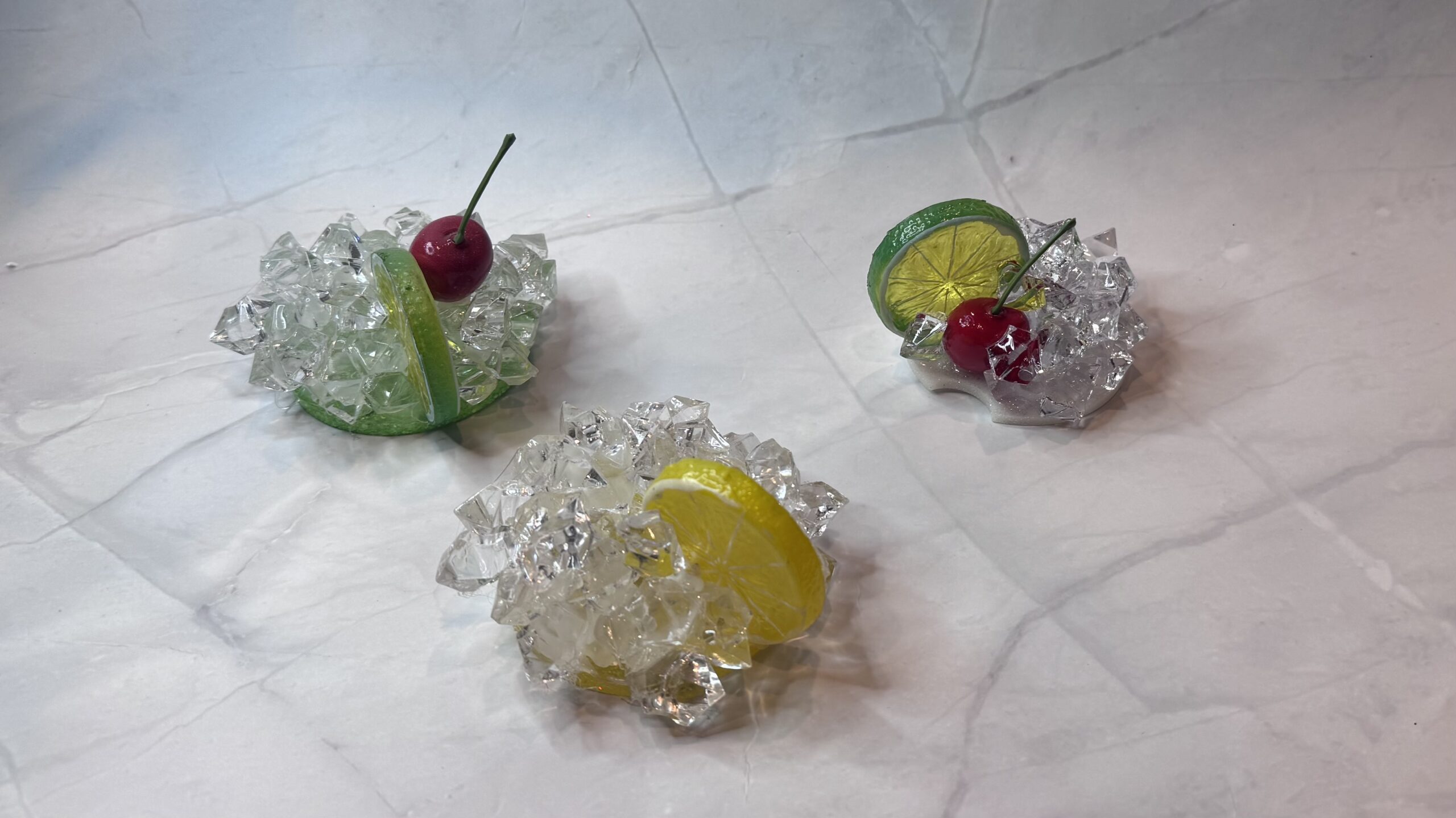 Three faux drink garnishes featuring cherries, lemon or lime slices, and sparkling ice pieces on marble.