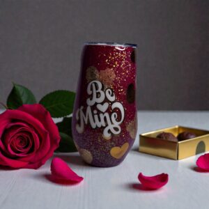 Glittery 'Be Mine' tumbler with gold polka dots, rose, and chocolates on white surface
